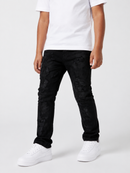 A person wearing black distressed skinny jeans with a subtle tonal pattern, styled with a white t-shirt and white sneakers against a light gray studio background.