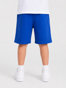 A child wearing bright royal blue athletic shorts, a white t-shirt, white socks, and white sneakers, shown from the waist down against a light gray studio background.