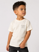 A young boy wearing a cream-colored short-sleeve t-shirt with a distressed texture and a gray 'MONEY DEPT. HUELLOCOD' graphic on the chest, paired with dark jeans, photographed against a light gray studio background.