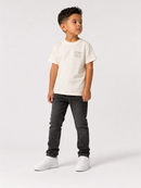 A young boy modeling a short-sleeve t-shirt with a distressed print, paired with dark gray jeans and white sneakers, standing against a light gray studio background.