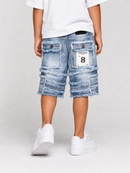 A child shown from the back wearing light blue frayed denim shorts with layered ruffle details, a white t-shirt, and white sneakers on a light gray studio background.