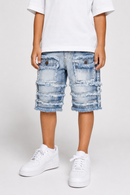 A child wearing light-wash blue distressed denim shorts with frayed edges and layered pocket details, styled with a white t-shirt and white sneakers against a light gray studio background.