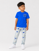 A young boy modeling a bright blue short-sleeve t-shirt with a blue graphic on the chest, paired with light-wash distressed jeans featuring blue ripped details, and white sneakers, set against a plain light gray studio background.