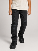 A person wearing black distressed jeans with ripped details and faded patches, paired with a light beige t-shirt and black sneakers, shown from the waist down on a light gray studio background.