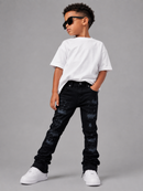 A young boy modeling a white crewneck t-shirt, black distressed jeans, and white sneakers, wearing black sunglasses, posed against a solid gray studio background.
