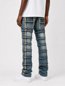 A person shown from the waist down wearing distressed blue denim jeans with patchwork paneling and frayed seam details, paired with a white t-shirt and white sneakers, against a light gray studio background.