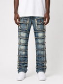 A person wearing blue denim jeans with distinctive horizontal patchwork paneling and frayed trim details, styled with a white t-shirt and white sneakers against a light gray studio background.