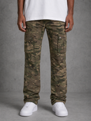 A person wearing camouflage cargo pants with multiple flap pockets, paired with a white t-shirt and white sneakers, standing against a textured gray studio background.