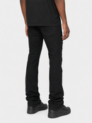 Rear view of a person wearing black jeans with distinctive vertical stitched detailing down the outer side of each leg, paired with a black t-shirt and black sneakers, against a light gray background.