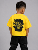 A child model shown from the back wearing a bright yellow t-shirt with a black graphic of a hand holding a puzzle piece and the text 'DON'T JUDGE ME' in yellow. The child is also wearing black jeans against a solid gray studio background.