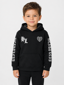 A young boy wearing a black pullover hoodie with white graphic prints on the chest and down both sleeves, paired with black pants, photographed against a light gray studio background.