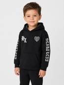 A young boy wearing a black pullover hoodie with white graphic prints on the chest and both sleeves, paired with black pants, photographed against a light gray studio background.