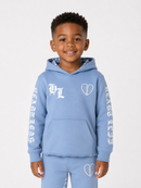 A young boy wearing a light blue hoodie and matching sweatpants set from Christian Dior Kids, featuring white gothic lettering and heart graphics, photographed against a light gray studio background.