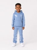 A young boy modeling a light blue two-piece tracksuit with white graphic prints, including a hoodie and matching sweatpants, paired with white sneakers, against a plain light gray background.