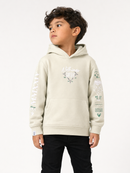 A young boy wearing a light beige Arkiwear hoodie with graphic prints on the chest and sleeves, paired with black pants, photographed against a plain white studio background.