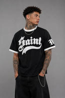 A man wearing a black ringer t-shirt with a white Gothic-style 'Saint' graphic on the chest and white trim on the collar and sleeves, paired with black pants, against a solid gray studio background. By Fresh N Fitted