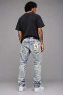 A man viewed from behind wearing light-wash, heavily distressed blue jeans with a cream patch on the back pocket, a black t-shirt, and white sneakers, standing against a gray studio background. By Fresh N Fitted