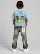A child model shown from the back wearing a light blue graphic t-shirt with 'TOP DOG 00' printed on the back, paired with distressed blue jeans and white sneakers, against a neutral gray studio background. By Fresh N Fitted.