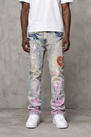 A person wearing distressed and painted light tint jeans with pink graffiti-style graphics, a white t-shirt, and white sneakers, standing against a textured gray concrete wall. By Fresh N Fitted.