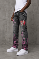 A person wearing washed black acid-wash jeans with pink flame and heart graphics, styled with a white t-shirt and white sneakers against a gray concrete wall. By Fresh N Fitted.