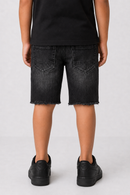 A child shown from the back wearing black denim shorts with a raw frayed hem, a black t-shirt, and black sneakers against a light gray studio background.