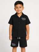 A young boy modeling a black short-sleeve button-up shirt and matching black shorts set, both featuring white script embroidery on the chest and left leg, against a plain light gray background.
