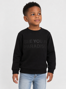A young boy wearing a black crewneck sweatshirt with the text 'SEE YOU IN PARADISE' printed in a dark gray tone on the chest, paired with light blue jeans, against a plain light gray background.
