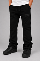 Black sweatpants with 'SEE YOU IN PARADISE.' printed in tonal black on the upper left thigh, shown on a model wearing a white t-shirt and black sneakers against a gray studio background.