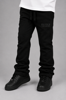 Black sweatpants with 'SEE YOU IN PARADISE' printed on the left thigh, worn with a white t-shirt and black sneakers against a gray studio background.
