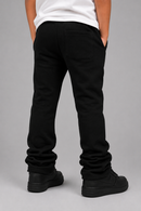 Rear view of a person wearing black sweatpants with a white t-shirt and black sneakers, standing against a solid gray studio background.