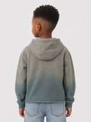 A child model shown from the back wearing a hooded sweatshirt with a vertical gradient color-block design, fading from a muted sage green at the top to a slate blue at the bottom, paired with light blue jeans against a plain light gray background.