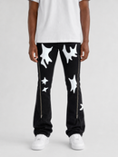 A model from the waist down wearing black pants with white abstract star-like patches and silver side zippers, paired with a white t-shirt and white sneakers, on a light gray studio background.