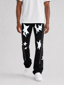 A person wearing black pants with large white abstract star-like patches and white sneakers, shown from the waist down against a light gray studio background.