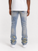 Rear view of a person wearing light-wash blue distressed jeans with yellowed abrasion details near the cuffs, paired with a white t-shirt and white sneakers on a light gray studio background.