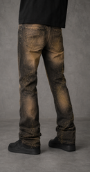 Rear view of a person wearing distressed brown and black jeans with metallic gold glitter details, paired with black shoes and a black top, against a gray studio background.