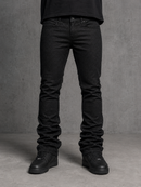 A person wearing slim-fit black jeans with black sneakers, shown from the waist down against a textured gray studio backdrop.