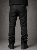 Rear view of a person wearing slim-fit black jeans with a black shirt and black shoes, standing against a textured gray studio background.