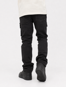 Rear view of a person wearing black cargo-style jeans with flap side pockets, a light cream top, and black sneakers against a plain light gray studio background.