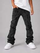A person modeling a pair of heavily distressed black denim jeans with raw frayed edges and patchwork paneling, styled with a white t-shirt and white sneakers against a neutral gray studio background.