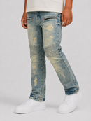 A person wearing light blue distressed denim jeans with faded yellowish patches and reinforced seam panels, paired with white sneakers against a light gray studio background.