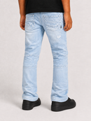 A person shown from the back wearing light-wash blue jeans with visible seam detailing and a small distressed patch near the upper right thigh, paired with a black shirt and black shoes on a light gray studio background.