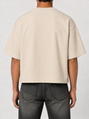 A person shown from the back wearing a plain short-sleeve cropped T-shirt in a light warm off-white/cream color, paired with dark gray jeans, against a light gray studio background.