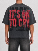 A person shown from the back wearing a faded black graphic T-shirt with large distressed red text reading “IT’S OK TO CRY” over a black graphic background, paired with dark jeans against a light gray studio backdrop.