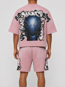 A model shown from the back wearing a light pink short-sleeve crop top and matching shorts set. The top features a large graphic print of a blue skull surrounded by black and white graffiti-style lettering.