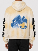 A person shown from the back wearing a hooded sweatshirt with a cream and yellow tie-dye wash, a large blue abstract graphic on the back, and black lettering down both sleeves, paired with black pants against a light gray studio background.