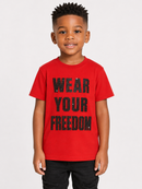 A young boy wearing a bright red t-shirt with 'WEAR YOUR FREEDOM' printed in black distressed letters, paired with black pants, against a light gray background.