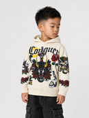 A young boy wearing a cream-colored Conquuer hoodie with a detailed black, red, and yellow graphic print on the front and sleeves, paired with dark cargo pants, against a light gray studio background.