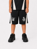 A child wearing black athletic shorts with white side stripes and white printed graphics, paired with black sneakers, shown against a light gray studio background.