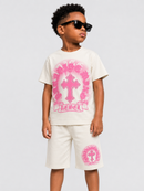 A young boy modeling a cream-colored two-piece outfit with a pink graphic print. The set includes a short-sleeve t-shirt and matching shorts, both featuring a pink circular design with a cross and the word 'RENE'.