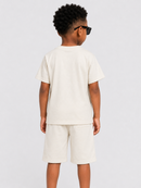 A child model shown from the back wearing a cream-colored short-sleeve t-shirt and matching shorts, styled with black sunglasses, against a light gray studio background.